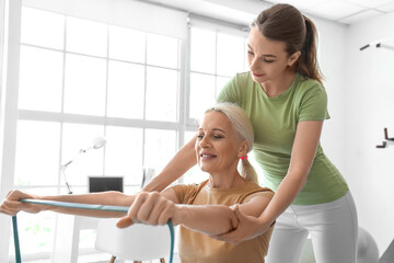 Fototapeta premium Mature woman training with elastic band and female physiotherapist in rehabilitation center