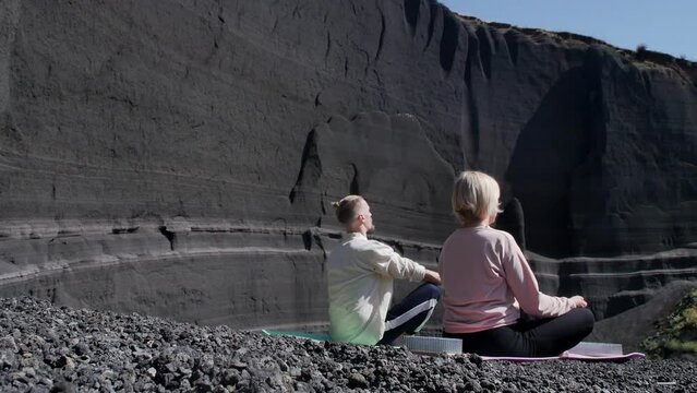 A view from behind two yogis, a man and a woman sitting in a lotus position on rugs, meditate in the crater of an extinct volcano with gray slag rocks. Meditation man and woman outdoors