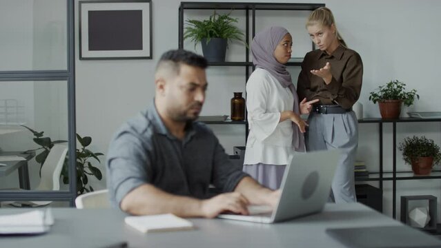 Selective focus shot of ignorant young woman standing with her colleague spreading gossips or defaming their Middle Eastern co-worker