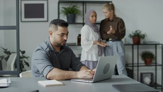 Selective focus shot of Middle Eastern man working on laptop in office trying to avoid his female colleagues talking nonsense about himSelective focus shot of Middle Eastern man working on laptop in o
