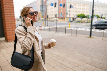 Stylish woman entrepreneur talking phone whiile standing on modern building background with coffee