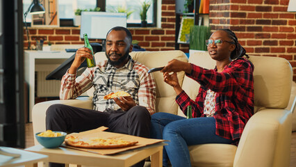 African american partners eating pizza slices at home, feeling happy watching film together. Relaxed couple having fun eating takeaway delivery food in living room, leisure activity.