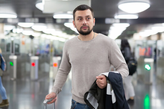 Positive young man going on trip, walking with suitcase to train in subway on blurred background of modern electronic turnstiles..