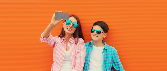 Happy smiling mother with son teenager taking selfie with smartphone on orange background