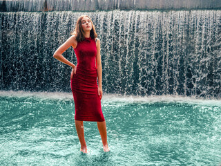 Teenager girl in high style red dress posing in water, waterfall behind her back. Prom photoshoot in beautiful location Young lady model ready for prom party. Confident person. Selective focus. © mark_gusev