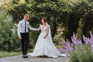 Fototapeta premium portrait of the bride and groom in the park, the bride and groom are walking holding hands near lavender flowers. Wedding walk in the park. Long train of the dress