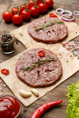 Raw burger patties on parchment and hamburger ingredients on a wooden background.