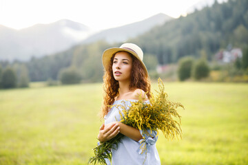 Woman with brown red hair raising hat against mountain and blooming flowers on windy day in countryside