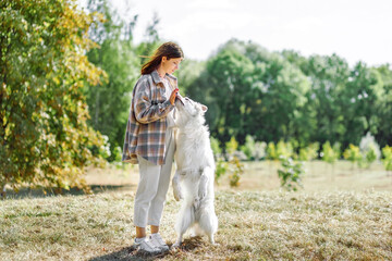 Stylish woman backpack training cute dog in sunny autumn woods. Teamwork. Young female hipster giving five to swiss shepherd white dog. Travel and hiking with pet.