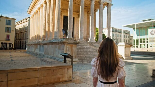 Young woman explores The Roman temple in Nimes, Occitanie, France on a sunny summer day. Beautiful girl in a dress looks at the Antique temple decorated with columns and friezes in France.
