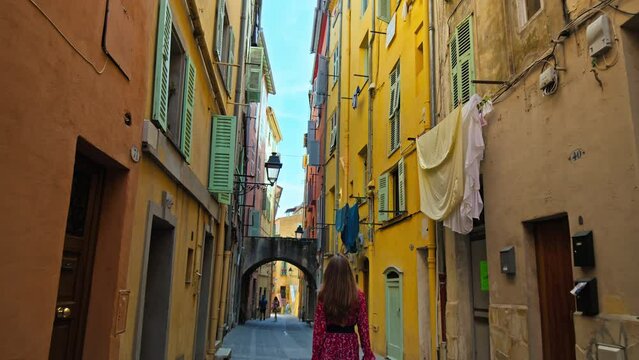 Cinematic view of a woman with a camera walking on a narrow street in Menton. Beautiful girl in a dress exploring French Riviera and enjoying the colorful houses in Menton.