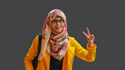 Close up portrait of smiling young Muslim woman wearing yellow jacket and brown hijab with red flower motif left hand making V sign of victory gesture. Happy and cheerful expression.