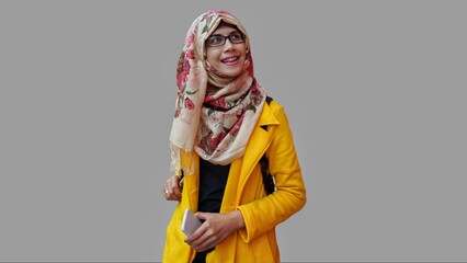 Close up portrait of smiling young Muslim woman wearing yellow jacket and brown hijab with red flower motif looking up while holding phone. Happy and self confidence expression.