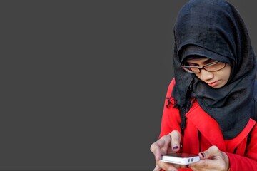 Close up portrait of young Muslim woman wearing black hijab and red jacket holding and looking at phone. Serious expression.