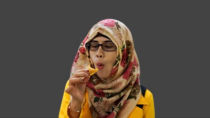 Close up portrait of smiling young Muslim woman wearing yellow jacket and brown hijab with red flower motif holding and eating small brown cake.