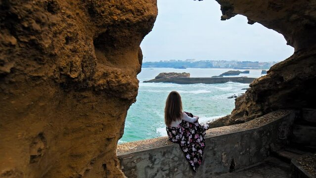 Elegant young girl sitting on a rocky window by seashore cliffs in Biarritz. A tourist woman at Rocher de la Vierge arch looking at blue waves of the sea enjoying the Basque coast in South of France.
