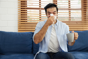 young man having a cold, using tissue paper and blowing his nose on sofa