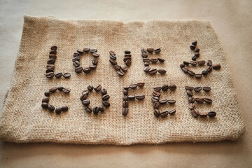 Inscription love coffee from coffee beans on burlap background. Banner for cafe or coffee shop advertising in rustic style. Roasted arabica on jute bag backdrop, close up.