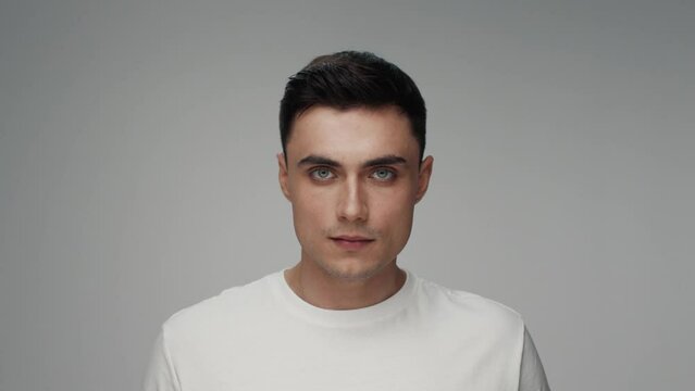 Portrait of Young Man Looking at Camera in Color Studio Shot. Adult Boy Isolated Alone on Grey Background Close-up. Beautiful 20s Person in Trendy t-shirt Raising Head and Opening Bright Pretty Eyes