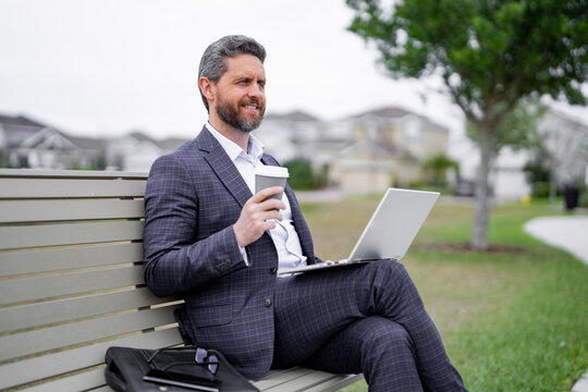 Business man sitting on bench in park, work on laptop, drink coffee. Businessman online work from laptop. Business man rest and reading news in american neighborhood. Business man is relax on bench.