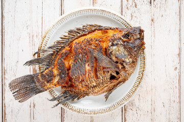 tasty large fried nile tilapia fish in circle ceramic plate on white wood texture background, top table view, flat lay