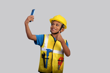 Portrait Indian Asian kid boy aged 7 to 8, wearing a yellow helmet and Safety jacket. Taking selfie with smartphone showing Victory with two fingers. He had a dream to future study as an engineer. He 