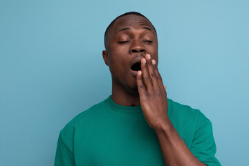 young american man in t-shirt yawns on blue background with copy space. people lifesal concept