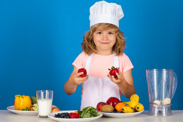Portrait of a 7, 8 years old child in cook cap and apron hold strawberries making fruit salad and cooking food in kitchen. Cute little blonde happy smiling chef.