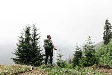 Portrait of a man in the mountains: he stands with a backpack on his back, in front of him, a view of a misty valley unfolds.