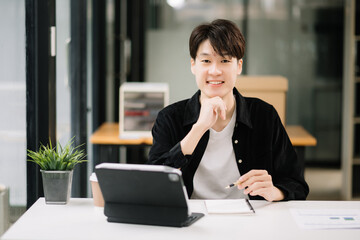 Fototapeta premium Young attractive Asian man smiling thinking planning writing in notebook, tablet and laptop working from home, looking at camera at office .