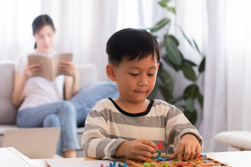 Asian Adorable kid boy playing wordplay from toys while waiting mother read a favorite book in the living room. son is focusing on a board game with word puzzles on the weekend.