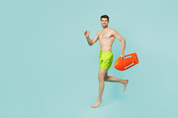 Full body side view strong young lifeguard man wear green shorts swimsuit relax near hotel pool hold lifebuoy walk go run isolated on plain blue background. Summer vacation sea rest sun tan concept.
