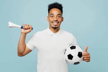 Happy smiling young man fan wears basic t-shirt cheer up support football sport team hold soccer ball blowing pipe show thumb up watch tv live stream isolated on plain pastel blue color background..