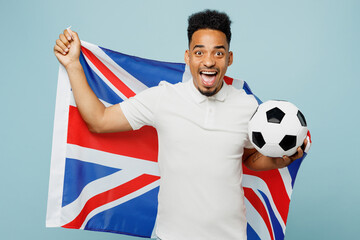 Young overjoyed excited happy fun man fan wearing basic t-shirt cheer up support football sport team hold British flag soccer ball watch tv live stream isolated on plain pastel blue color background.