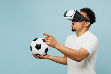 Young shocked man fan wearing basic t-shirt cheer up support football sport team hold in hand soccer ball watching in vr headset pc gadget watch tv live stream isolated on plain blue color background.