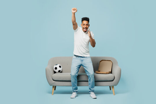 Full body expressive young man fan wear t-shirt cheer up support football sport team hold soccer ball sit on grey sofa do winner gesture scream watch tv live stream isolated on plain blue background.