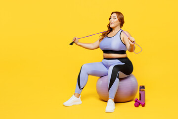 Fototapeta na wymiar Full body young chubby plus size big fat fit woman wear blue top warm up training sit on fit ball hold in hand skipping rope isolated on plain yellow background studio home gym. Workout sport concept.