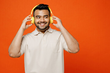 Young smiling happy cheerful Indian man wears white t-shirt casual clothes listen to music in headphones look aside on area isolated on plain orange red background studio portrait. Lifestyle concept.