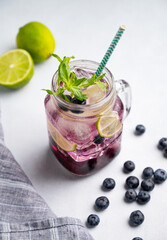 Blueberry mojito or lemonade with lime, ice and mint in a glass on a light background with berries and fruit close up. The concept of a summer refreshing drink