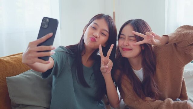 
Two young smiling asian women taking selfie self portrait photos on smartphone in living room at home. Happy lifestyle.