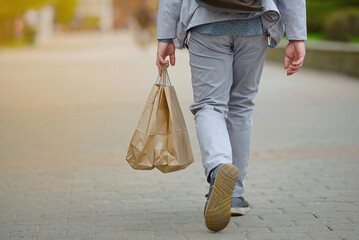 Man holding paper shopping bag walking down the street, rear view. Man shopaholic walking alone. - obrazy, fototapety, plakaty