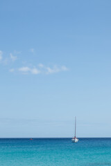 Fototapeta premium Seascape. Turquoise blue sea with sailboat in the background, sky with white clouds. Fuerteventura, Canary Islands, Spain 