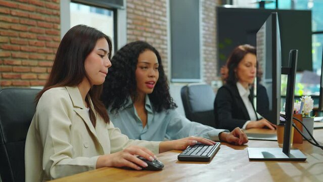 Two young women use computers to work at the office. They discuss projects together in the office.