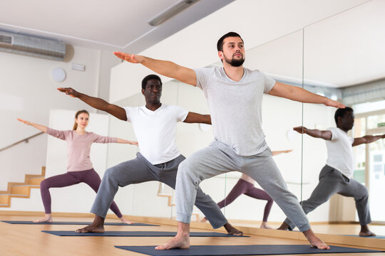 Caucasian man practising warrior II pose with people during group yoga training.