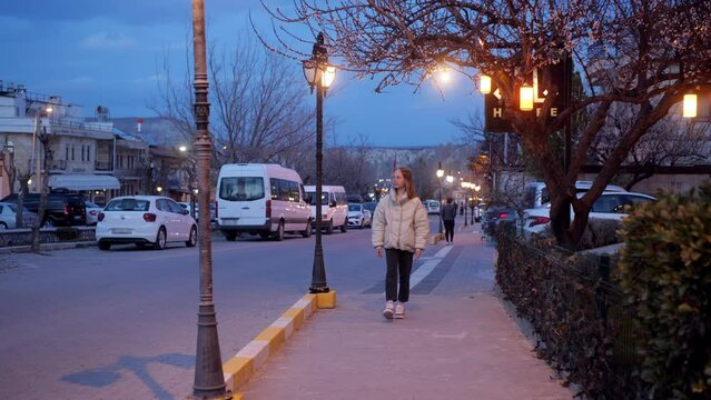 Blonde teen girl enjoying walking down street with streetlights in evening in city. Calm girl in jacket looking around going on sidewalk. Leisure, pastime, urban lifestyle concept. Teenager strolling.