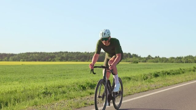 Active caucasian male in helmet and sunglasses biking on even roadway along summer field in early evening, Confident rider making cycling experience more enjoyable while observing landscapes.