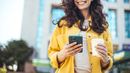 Modern young woman walking on the city street texting and holding cup of coffee. Smiling woman using mobile phone while having a coffee. Business woman texting on her phone