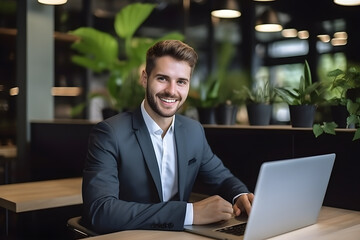 Smiling young businessman working laptop in modern office on colleagues background. Professional entrepreneur sitting in front of laptop, smiling at camera, copy space.