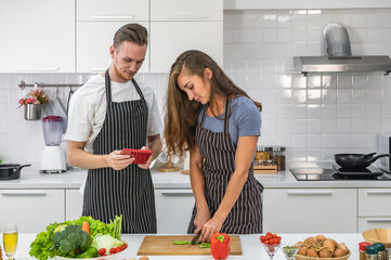 Caucasian young couple in white kitchen cooking making preparing healthy food with vegetables wearing aprons holding a tablet device.