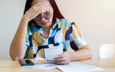 Frustrated Asian woman holding and looking at bills and calculating her monthly expenses and debts...